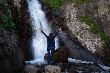 Şelaleyi süsleyen genç ve mutlu bir kadın. Şelalenin önünde duran, elleri kırışmış ve gülümseyen bir kadın. Çevre seyahati kavramı. Şelale Shumka, Dombai, Karachay-Çerkessia, Rusya