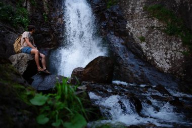 Sırt çantalı yürüyüşçü waalkinh güzel doğa manzarasında parktaki şelaleye bakıyor. Açık havada duran yetişkin erkek portresi. Şelale Shumka, Dombai, Rusya
