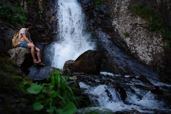 Sırt çantalı yürüyüşçü waalkinh güzel doğa manzarasında parktaki şelaleye bakıyor. Açık havada duran yetişkin erkek portresi. Şelale Shumka, Dombai, Rusya