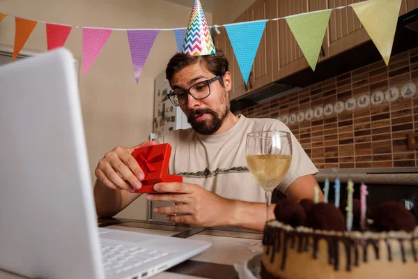 Man celebrating birthday online in quarantine time. The guy opens the box and is very happy with the gift. Communicating with friends remotely