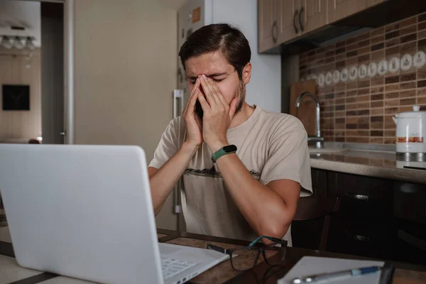 Tired young man feel pain eyestrain holding glasses rubbing dry irritated eyes fatigued from computer work, stressed man suffer from headache bad vision sight problem sit at home table using laptop