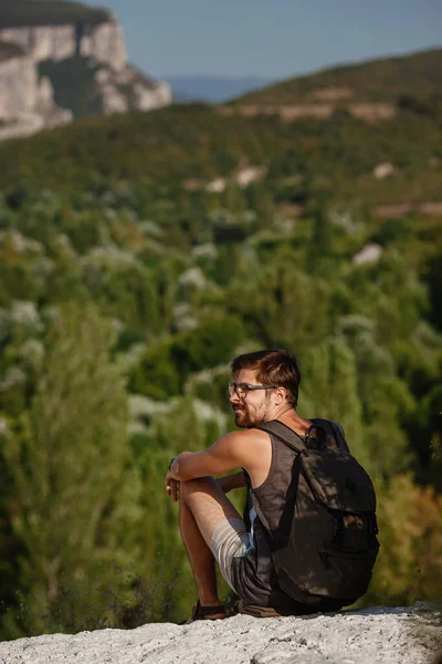 Sakin yaz günbatımında sırt çantasıyla dağın tepesinde dinlenen genç bir erkek yürüyüşçü. Tatilden kalma manzara. Yorgun gezgin adam tepede dinleniyor..