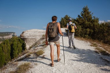 İki adam kayalık yamaçlarda yürüyüş çubuklarını kullanarak sırt çantalarını taşıyorlar. Dağlar Vadisi, insanların yanı sıra manzaralı. Doğaya yaz seyahati. Arka planda yürüyüşçü.