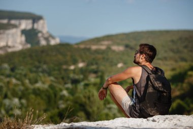 Sakin yaz günbatımında sırt çantasıyla dağın tepesinde dinlenen genç bir erkek yürüyüşçü. Tatilden kalma manzara. Yorgun gezgin adam tepede dinleniyor..