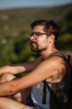 Sakin yaz günbatımında sırt çantasıyla dağın tepesinde dinlenen genç bir erkek yürüyüşçü. Tatilden kalma manzara. Yorgun gezgin adam tepede dinleniyor..