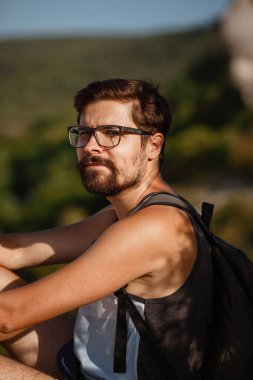 Sakin yaz günbatımında sırt çantasıyla dağın tepesinde dinlenen genç bir erkek yürüyüşçü. Tatilden kalma manzara. Yorgun gezgin adam tepede dinleniyor..