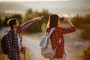 Yürüyüş gezilerinde eğlenen genç çiftin portresi. Kafkasyalı ve Asyalı yürüyüşçü çift yaz tatilinde eğleniyorlar. Doğayı dürbünle izliyorlar..