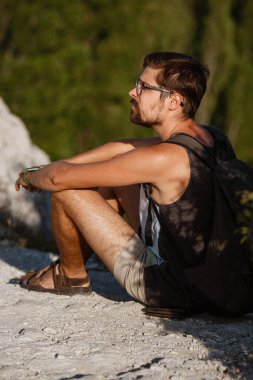 Sakin yaz günbatımında sırt çantasıyla dağın tepesinde dinlenen genç bir erkek yürüyüşçü. Tatilden kalma manzara. Yorgun gezgin adam tepede dinleniyor..