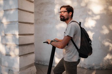 Şehirde elektrikli scooter sürmek eğlenceli. Hipster bir adam yaz günbatımında şehirde yürümekten hoşlanır. Fikir ve konsept ekoloji, stil ve eğlence şehirde dolaşıyor.