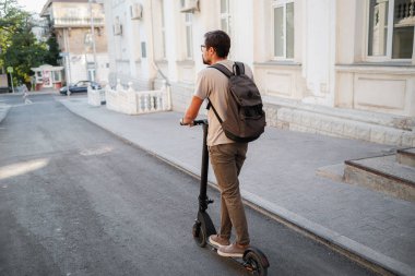 Şehirde elektrikli scooter sürmek eğlenceli. Hipster bir adam yaz günbatımında şehirde yürümekten hoşlanır. Fikir ve konsept ekoloji, stil ve eğlence şehirde dolaşıyor.