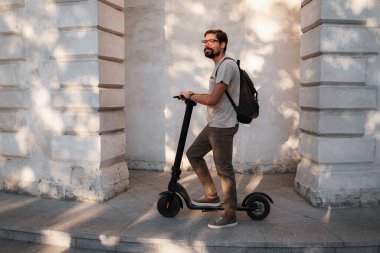 Şehirde elektrikli scooter sürmek eğlenceli. Hipster bir adam yaz günbatımında şehirde yürümekten hoşlanır. Fikir ve konsept ekoloji, stil ve eğlence şehirde dolaşıyor.
