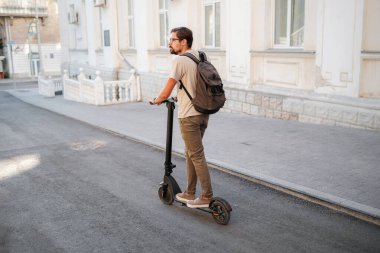 Şehirde elektrikli scooter sürmek eğlenceli. Hipster bir adam yaz günbatımında şehirde yürümekten hoşlanır. Fikir ve konsept ekoloji, stil ve eğlence şehirde dolaşıyor.