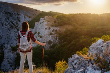 Sırt çantalı ve sırıklı formda bir kadın yürüyüşçü kayalık bir dağ sırtında dikiliyor ve sağlıklı bir doğa yaşam tarzı kavramının zirvesine çıkıyor.
