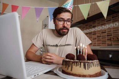 Karantina zamanında doğum gününü kutlayan bir adam. Adam doğum gününü kutluyor, pastadaki mumu söndürüyor ve video görüşmesi yapıyor. Coronavirüs salgını 2020.