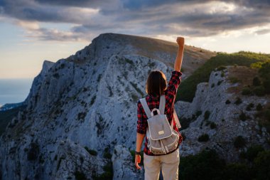 Sırt çantalı formda bir kadın yürüyüşçü. Ellerini kaldırmış kayalık bir dağ sırtına bakıyor ve sağlıklı bir doğa yaşam tarzı kavramının zirvesine çıkıyor.