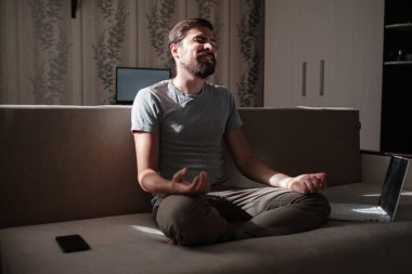 Milenyum hippi erkeği evdeki kanepede rahatça dinlenemiyor. Erkekler kanepede dizüstü bilgisayarla ve telefonla meditasyon yaparlar.