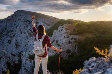 Sırt çantalı ve direkli formda bir kadın yürüyüşçü kayalık bir dağ sırtında ayakta duruyor ve sağlıklı bir doğa yaşam tarzının zirvelerine bakıyor.