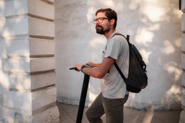 Şehirde elektrikli scooter sürmek eğlenceli. Hipster bir adam yaz günbatımında şehirde yürümekten hoşlanır. Fikir ve konsept ekoloji, stil ve eğlence şehirde dolaşıyor.
