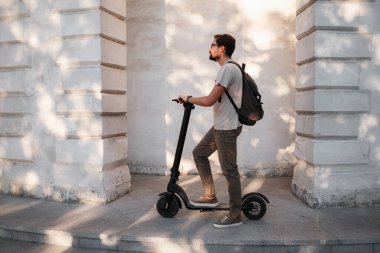 Şehirde elektrikli scooter sürmek eğlenceli. Hipster bir adam yaz günbatımında şehirde yürümekten hoşlanır. Fikir ve konsept ekoloji, stil ve eğlence şehirde dolaşıyor.