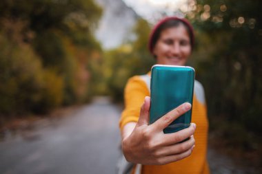 Sırt çantalı hippi kadın ormanda akıllı telefon cihazını tutuyor. Gezgin, dağ yolunun arka planında cep telefonu kullanıyor. sonbahar yaşam tarzı