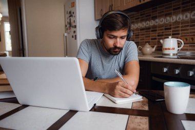 Odaklanmış genç adam internetten kulaklık takıyor laptopta webinar podcast izliyor eğitim konferansını dinliyor notlar alıyor çalışma masasında oturuyor, konsept öğreniyor.