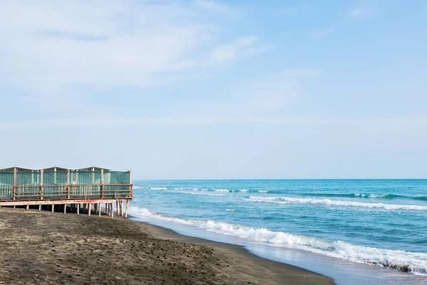 Piękne morze, czarna piaszczysta plaża i białe i niebieskie pasiaste domy plażowe — Zdjęcie stockowe