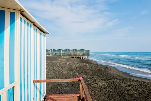 Beautiful sea, the black sandy beach and white and blue striped beach houses — Stock Photo, Image