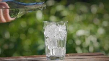 Yeşil yaz bahçesi arka planında, ahşap masadaki buzla birlikte, taze maden suyu bardağa dökülüyor. Yiyecek ve içecek yakın mesafede.