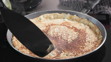 Aşçı, mutfakta spatulayla taze pişmiş krepi sıcak tavadan kaldırır. Sağlıklı kahvaltı için evde leziz krep yapma süreci, yiyecek kapanıyor.