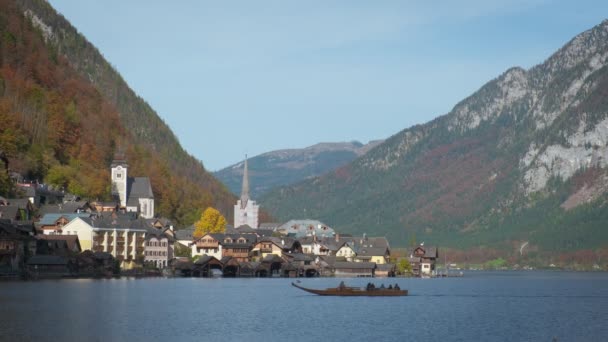 有名な観光地で秋の色オーストリアの山アルプスの牧歌的な町ハルシュタット 大聖堂教会 地元の家 高い丘の上の木の上の黄金の葉 湖に浮かぶボート — ストック動画