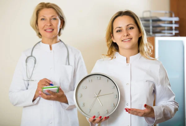 Dos médicos amigables sosteniendo el reloj y sonriendo en la clínica moderna —  Fotos de Stock