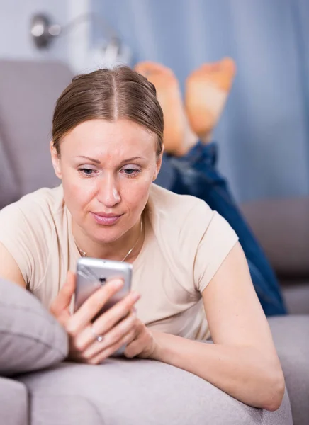 烦躁不安的女人躺在沙发上 通过电话阅读短信 — 图库照片