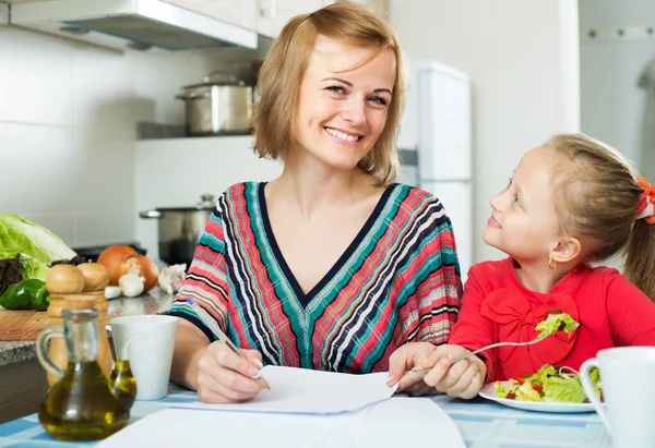 Mutfakta Çalışan Pozitif Bir Anne Yakınlarda Yemek Yiyen Küçük Bir — Stok fotoğraf