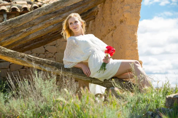 Junge Frau in weißem Kleid sitzt auf Baum und hält Mohnpflanzen in der Hand — Stockfoto