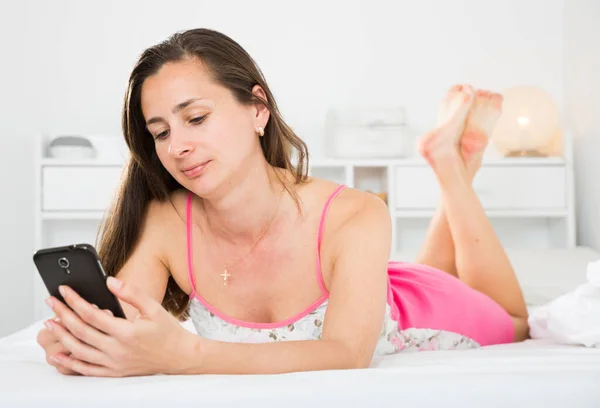 Adult positive woman using her phone while lying in bed — Stock Photo, Image