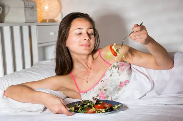 Kvinnan i nattlinne kappa äter sallad i sängen — Stockfoto