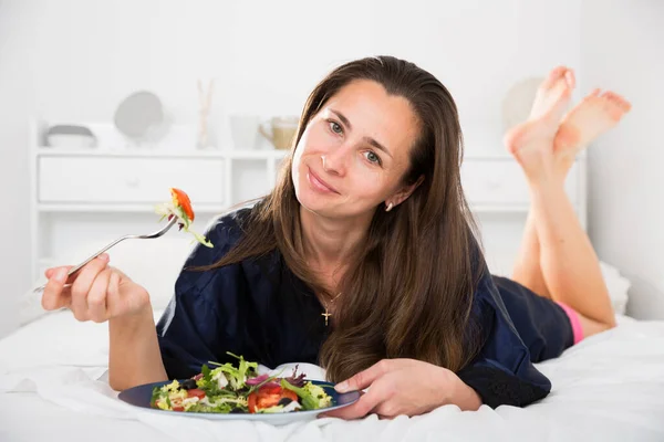 Ung vacker kvinna äter grönsakssallad från platon i sängen — Stockfoto