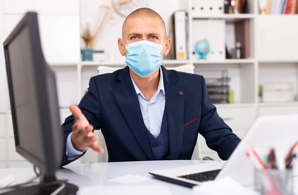 Hombre con máscara médica sentado en el escritorio con el ordenador portátil en la oficina —  Fotos de Stock