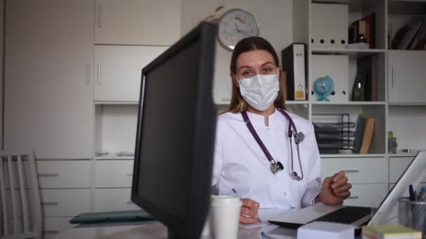 Doctora en bata de laboratorio y máscaras médicas protectoras hablando en el consultorio — Vídeos de Stock