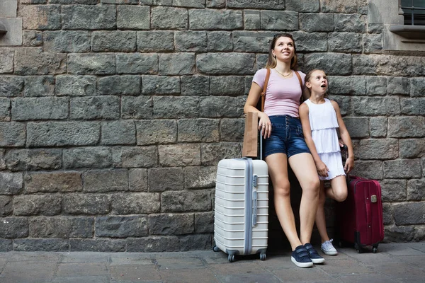 Женщина и маленькая девочка путешественница с чемоданом, прислонившись к каменной стене — стоковое фото