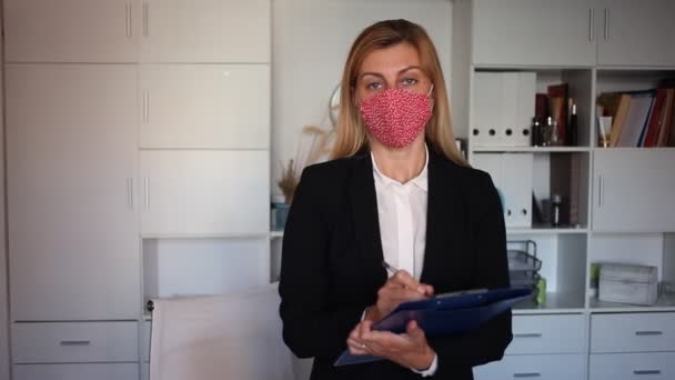 Молодая деловая женщина в маске для лица, стоящая с буфером обмена и вежливо приветствующая современный офис компании — стоковое видео