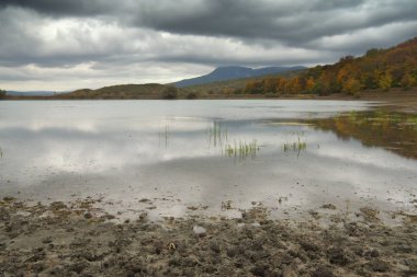 Sonbahar ağaçları ile tepelik dağlar arasında göl kenarında yağmur geliyor. Kir ve su birikintisi. Doğa kompozisyon.