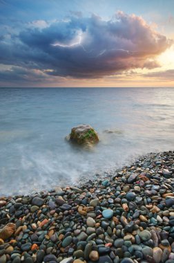 Deniz kıyısında gün batımı. Bulutlu gökyüzünde şimşek çakması. Taşlar ve yağmurlu bulut. Doğa yapısı bileşimi. 