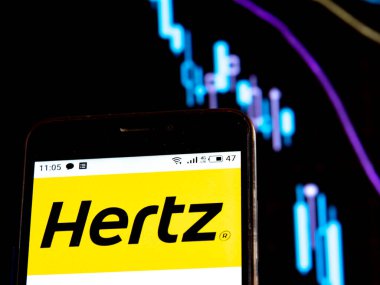 Bu resimde Hertz logosu bir grafiğin arka planında düşen eğrilerle birlikte bir akıllı telefonda görüntülenir. Coronavirus salgını sırasındaki uluslararası basında çıkan haberlere göre COVID-2019 En Büyük Araba Kiralama Şirketi Hertz