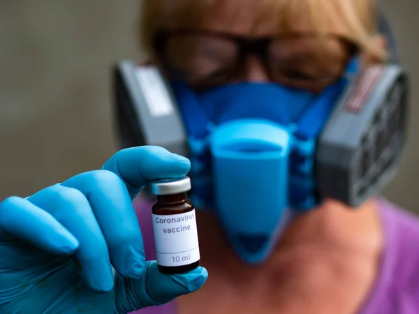 この写真では 灰色の背景に医療用マスクとゴム手袋を着用している科学者は 世界がコロナウイルスに対するワクチンを心配そうに待っているとして表示されたバイアルラベル付きコロナウイルスワクチンを示しています — ストック写真