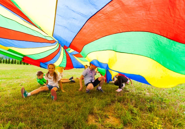 Crianças felizes escondidas sob paraquedas coloridos — Fotografia de Stock