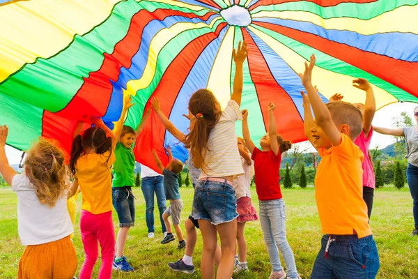 Joyous classmates jumping under colorful parachute in the summer outdoors — Stock Photo, Image