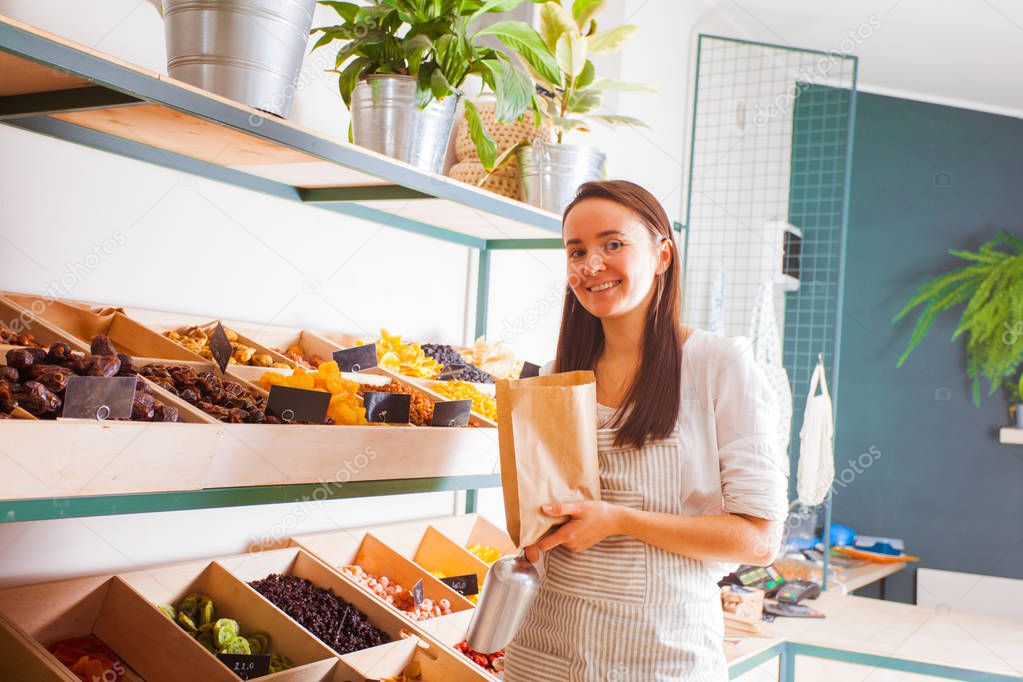 Pretty woman seller in the grocery shop, view of assortment of dry apricots, raisins, pears, dates, figs laying out on the rack with wooden boxes