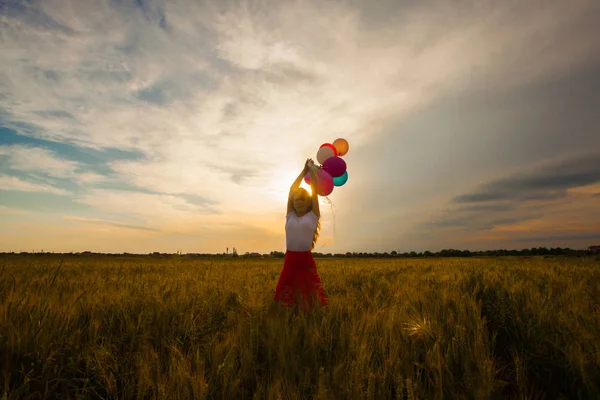 Menina com balões no campo de trigo e luz do sol no pôr do sol — Fotografia de Stock