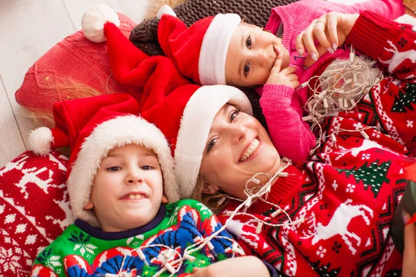 Вид сверху на счастливую мать с дочерью и сыном в Санта-Клаусах, лежа на полу дома — стоковое фото